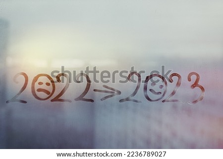 Handwritten blue arabic numbers 2022 with doodle sad face, arrow and date 2023 with smiling face on misted glass on window flooded with raindrops on background