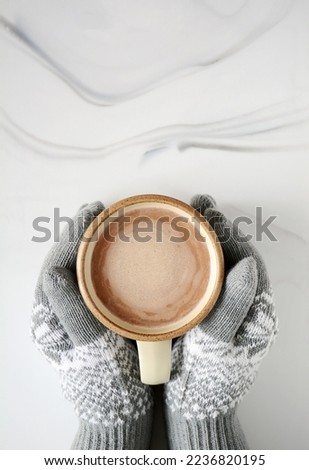 Mug of delicious hot chocolate in mug on marble background shot from overhead with room for text.  Seasonal comforting beverage hygge concept.  Hands in gloves holding mug.
