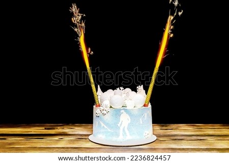 Lighting firework candle, sparkler on fire in blue sponge vanilla creamy cake with round marshmallow, chocolate snowflakes, fir trees, mountain, skier picture, sweet dessert on wooden table.