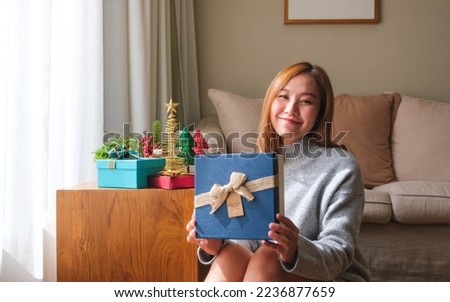 Portrait image of a young woman in sweater holding and showing a present box with Christmas holiday decoration at home