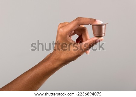 Crop anonymous African American female demonstrating small brown plastic coffee capsule on gray background