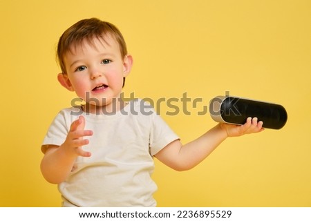 Toddler baby plays with a wireless music speaker on a studio yellow background. Happy child in a white t-shirt and blue jeans listens to music in an audio speaker. Kid aged one year four months