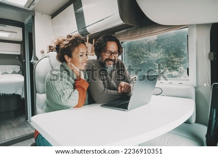 Cheerful couple using laptop together inside a camper van alternative house. Man and woman surfing the web or working on computer sitting and relaxing in indoor leisure activity during rv travel Royalty-Free Stock Photo #2236910351