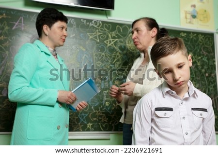 Sad upset schoolboy against the background of talking mom and teacher. Problems at school. The teacher complains to the mother about the student. Student with bad behavior at school. Royalty-Free Stock Photo #2236921691