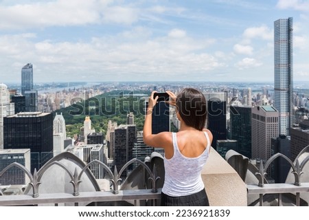 USA travel tourist in New York City vacation- woman looking at view of skyline taking photos with mobile phone from skyscraper. Girl traveling summer holidays United States road trip