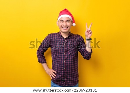 Cheerful young Asian man in Santa hat showing victory sign, looking at camera on yellow studio background. Happy New Year 2023 celebration merry holiday concept