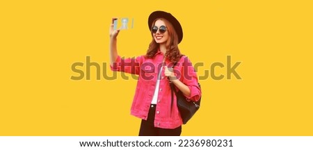 Portrait of happy smiling young woman taking selfie with smartphone wearing pink jacket, black round hat on yellow background