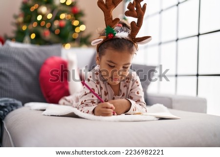Adorable hispanic girl drawing on notebook lying on sofa by christmas tree at home
