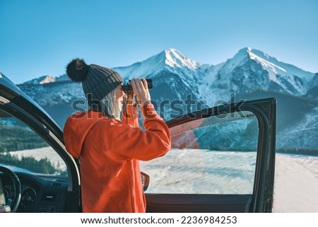 Woman traveling exploring, enjoying the view of the mountains, landscape, lifestyle concept winter vacation outdoors. Female standing near the car in sunny day, Girl looking through binoculars.