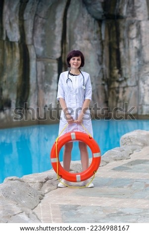 A brunette doctor in a white coat with a lifebuoy near the pool on the hotel grounds.