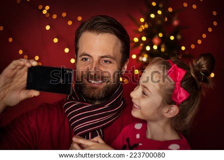 Christmas eve - smiling father and daughter taking photo of themselves (selfie) on mobile phone. Happy family time, christmas tree with lights on dark red as background.