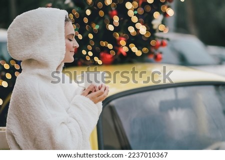 Beautiful young woman wearing white sweater with a hood drinking hot chocolate near yellow car with Christmas tree on the top. Happy winter holidays.