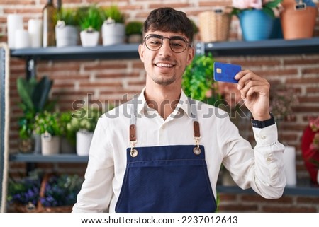 Young hispanic man working at florist shop holding credit card looking positive and happy standing and smiling with a confident smile showing teeth 