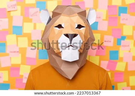 Photo of anonymous man wearing authentic outfit, creative lion paper mask, standing looking at camera, posing isolated over yellow background, stands against wall covered with memo cards.