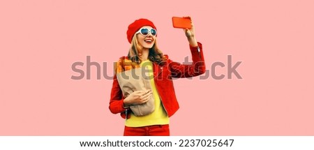 Portrait stylish happy smiling young woman taking selfie with smartphone holding grocery shopping paper bag with long white loaf bread wearing red beret, heart shaped sunglasses on pink background