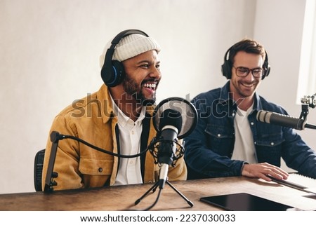 Happy radio presenter smiling while speaking into a microphone in a studio. Cheerful young man co-hosting an audio broadcast with a guest. Two young content creators recording an internet podcast. Royalty-Free Stock Photo #2237030033