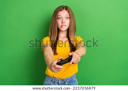 Photo of brown hair teenager schoolgirl wear yellow t-shirt bite lip nervous play video game wireless gamepad isolated on green color background
