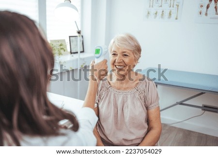 Adult female doctor checking temperature with contactless thermometer to sad senior lady patient in clinic office interior. Medical health care, exam, treatment illness during covid-19 pandemic
