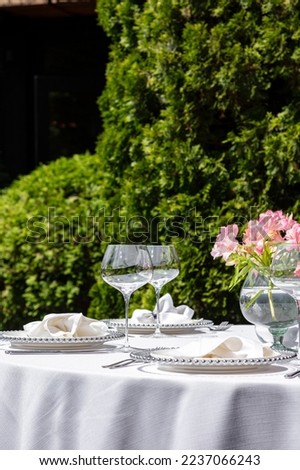 A round table with a white tablecloth stands in the middle of a lawn and a fence of grapes. Plates with underplates are served on the table. cutlery and wine glasses. There are chairs around the table