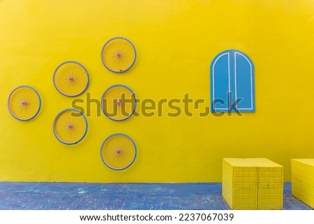 Yellow plain colour wall with blue colour window and bicycle rim. Selective focus with negative space. Royalty-Free Stock Photo #2237067039