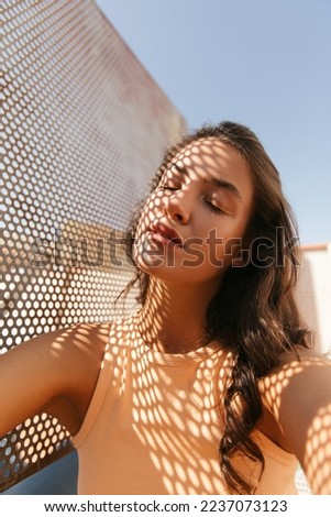 Close-up vertical image young caucasian woman relaxed with closed eyes outdoor in warm weather. Brunette wavy hair wears peach color top. Enjoying life concept.