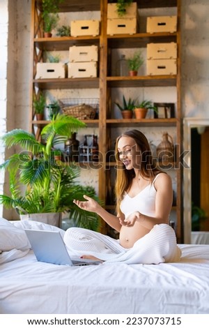 Photo of a pregnant woman sits at home in front of a laptop monitor - online consultation with a doctor or online meeting with friends. Woman waving hand and smiling. Vertical photo.