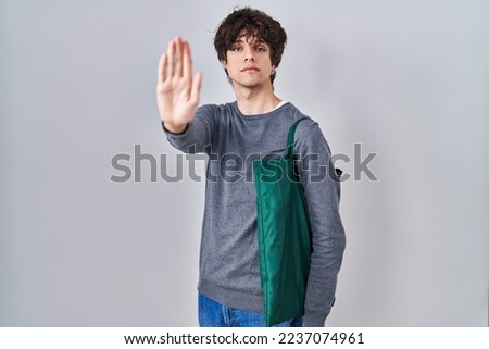 Young man wearing reusable bag with open hand doing stop sign with serious and confident expression, defense gesture 