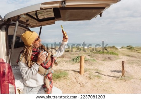 happy woman holding a mug leaning against a van next to beach and taking a selfie