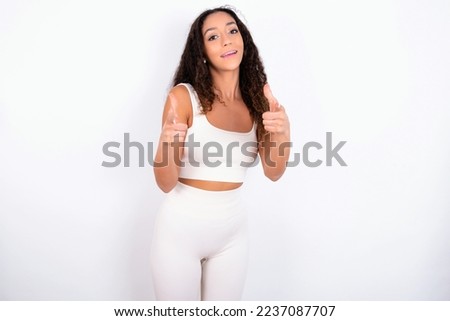 Beautiful teen girl with curly hair wearing white sport set over white background directs fingers at camera selects someone. I recommend you. Best choice