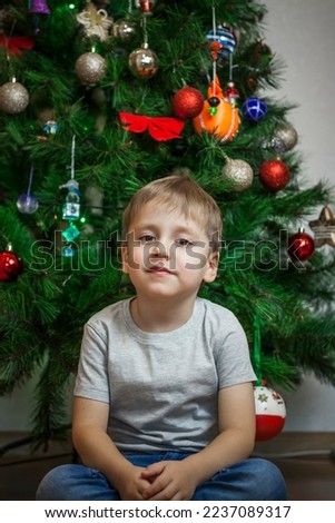 A photo of a beautiful boy in a gray T-shirt at the Christmas tree, looking into the camera. Portrait in a bright room. Natural, not staged photography. Children