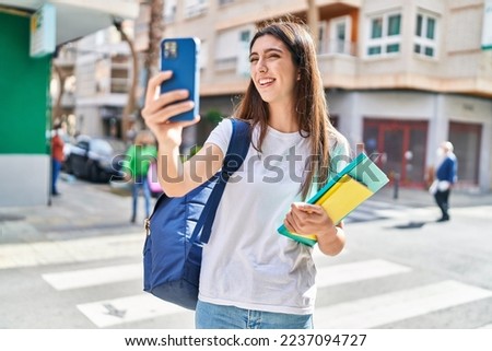 Young beautiful hispanic woman student holding books make selfie by smartphone at street