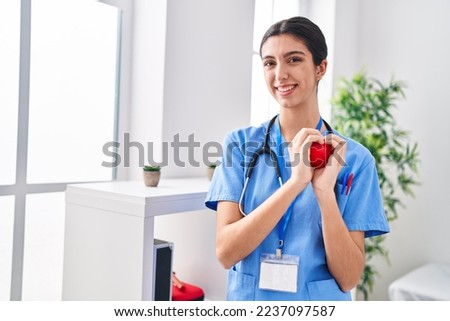 Young beautiful hispanic woman doctor smiling confident holding heart at clinic