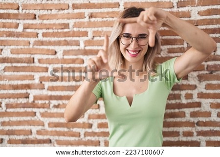 Young beautiful woman standing over bricks wall smiling making frame with hands and fingers with happy face. creativity and photography concept. 