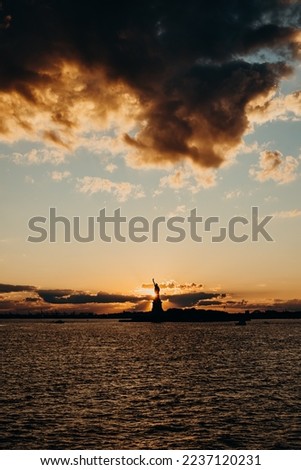 The picture shows the Statue of Liberty at sunset, with a warm glow over its copper surface and a colorful sky. The water surrounding the statue is calm. 