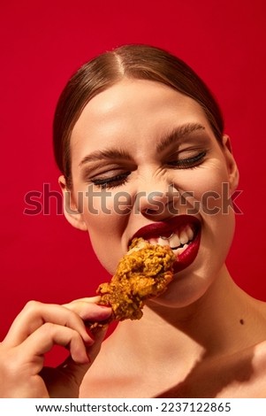 Young woman with red lipstick eating fried chicken, nuggets over vivid red background. Fast food lover. Food pop art photography. Complementary colors. Junk food. Copy space for ad, text