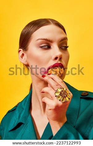 Young woman in green coat with red lipstick smudge, eating fried chicken, nuggets over yellow background. Fast food. Food pop art photography. Complementary colors. Copy space for ad, text