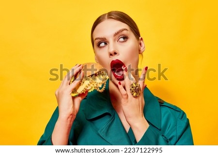 Young beautiful woman in green coat with red lipstick eating cheeseburger with necklaces over yellow background. Food pop art photography. Complementary colors. Copy space for ad, text