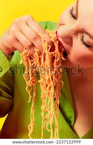 Close-up image of emotional young girl eating spaghetti, noodles with hands over yellow background. Feeling hungry, delicious taste. Food pop art photography. Complementary colors. Copy space for ad