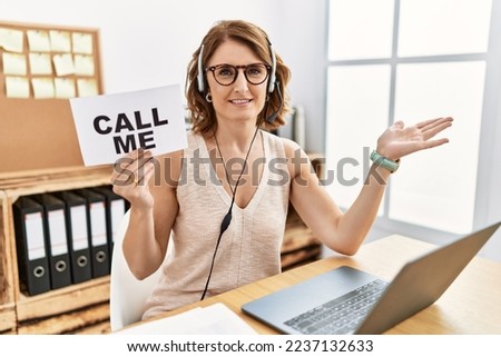 Middle age brunette woman wearing operator headset holding call me banner smiling cheerful presenting and pointing with palm of hand looking at the camera. 