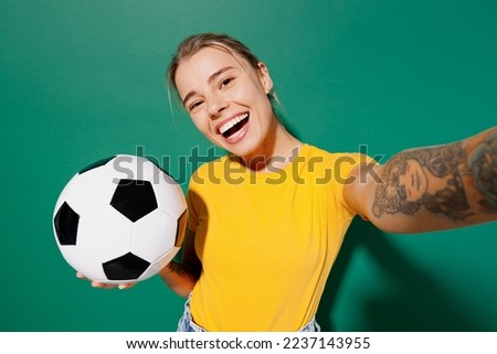 Young woman fan wear basic yellow t-shirt cheer up support football sport team hold soccer ball watch tv live stream doing selfie shot pov on mobile cell phone isolated on dark green background studio