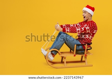 Full body satisfied merry fun young man 20s wearing red knitted Christmas sweater Santa hat posing sit ride sled isolated on plain yellow background. Happy New Year 2023 celebration holiday concept