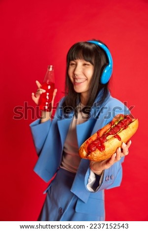 Unfocused portrait of young girl in stylish blue costume posing with hotdog isolated over red background. Street food. Concept of youth, beauty, fashion, lifestyle, emotions, facial expression. Ad