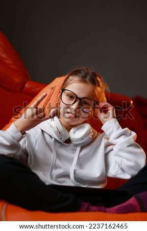 Attractive stylish little girl child wearing a casual sweatshirt and headphones, showing rock n roll sign and touching her glasses sitting in chair and looking in camera. Childhood lifestyle concept