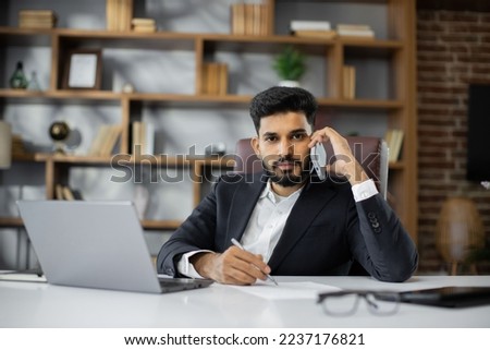 Successful young entrepreneur with attentive and concentrated look while having phone call, sitting at desk in modern office.