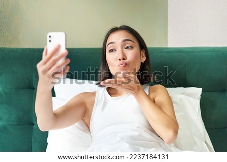 Beautiful asian girl lying in bed, making morning selfie, taking picture on smartphone in bedroom, smiling happily.