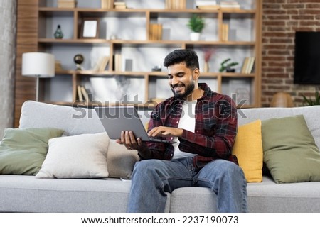 Bearded young man using laptop, sitting on couch at home, handsome guy having video call online in social network, having fun, watching movie, freelancer working on computer project