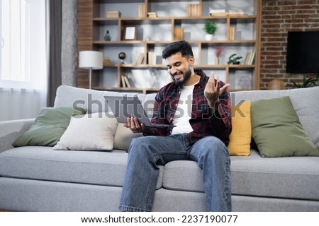 Young bearded businessman in checkered red shirt working on laptop computer, talking online while sitting on the comfortable sofa at home. Concept of remote work from home.