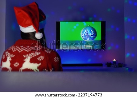 A woman sitting on sofa in a Christmas outfit looking at the TV (Christmas ball). Dressed in a Christmas sweater with reindeers and a Santa hat. All in blue tones and Christmas lights + candle.  