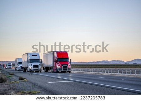 Convoy of a industrial grade commercial professional transportation of different big rig semi trucks with semi trailers driving on the straight highway road at twilight time in California Royalty-Free Stock Photo #2237205783