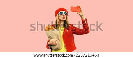 Portrait of stylish young woman taking selfie with smartphone holding grocery shopping paper bag with long white loaf bread wearing red beret, heart shaped sunglasses on pink background